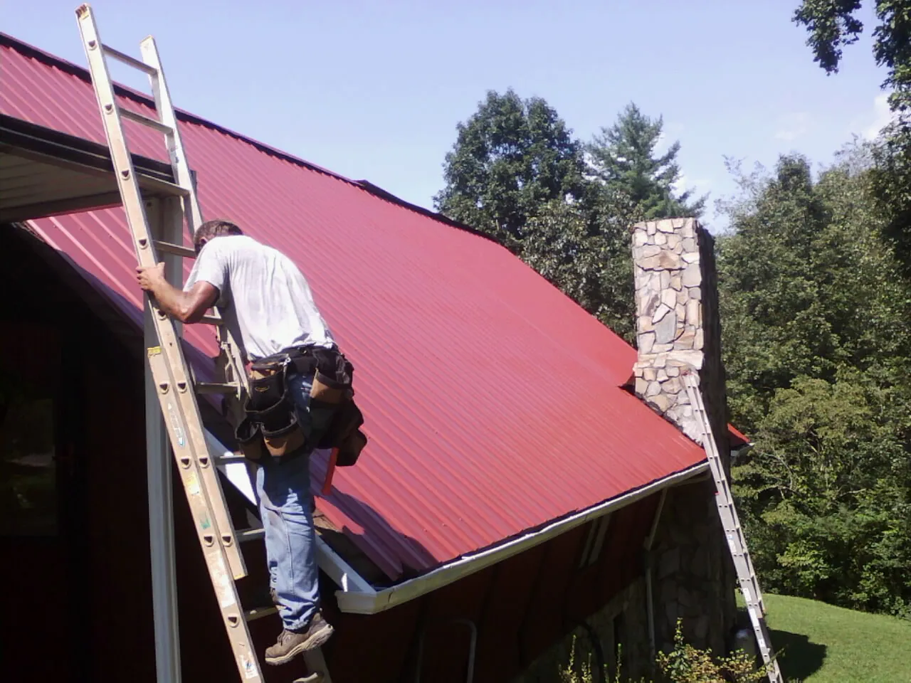 Roofer on a ladder beside a red metal roof and stone chimney — Soffit & Fascia Repair by Roofing Brothers in Malvern